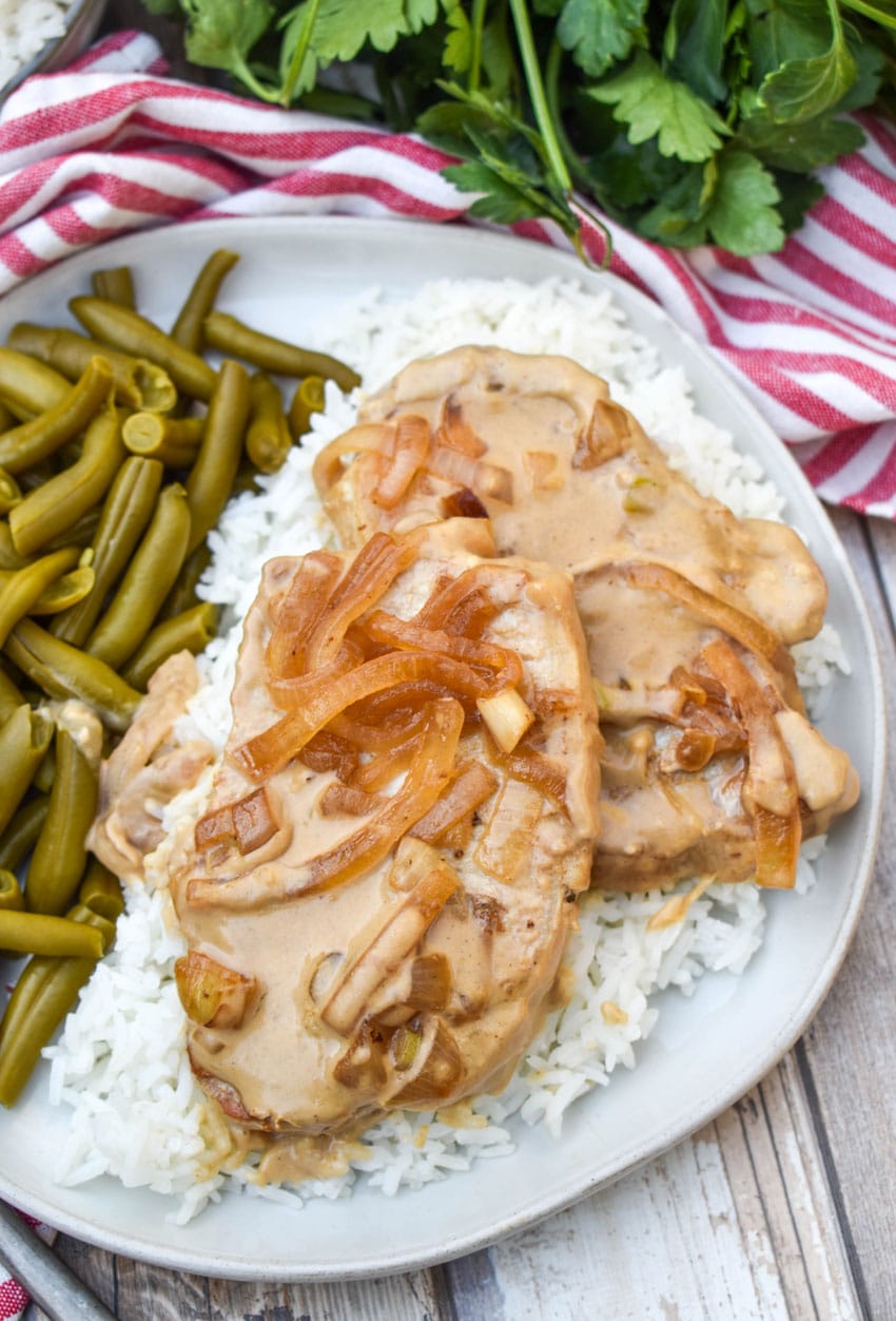 pork chops with cream sauce and onions over steamed white rice next to green beans on a white dinner plate
