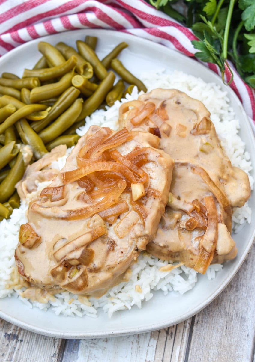 pork chops with cream sauce and onions over steamed white rice next to green beans on a white dinner plate