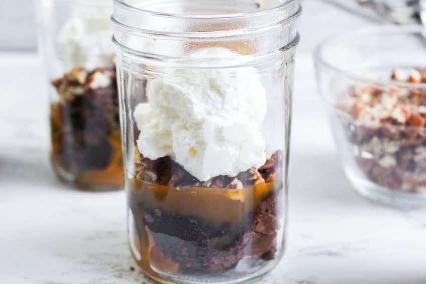 layers of brownie bits, caramel syrup, and chopped pecans topped with freshly whipped cream in small glass jars