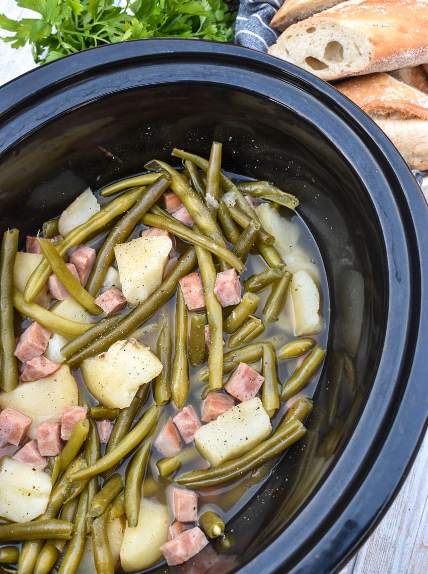 tender green beans with potatoes and chunks of ham in the black bowl of a slow cooker