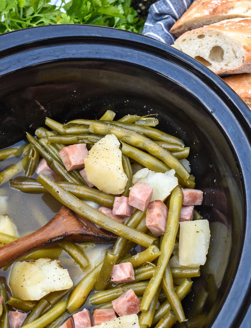 tender green beans with potatoes and chunks of ham in the black bowl of a slow cooker