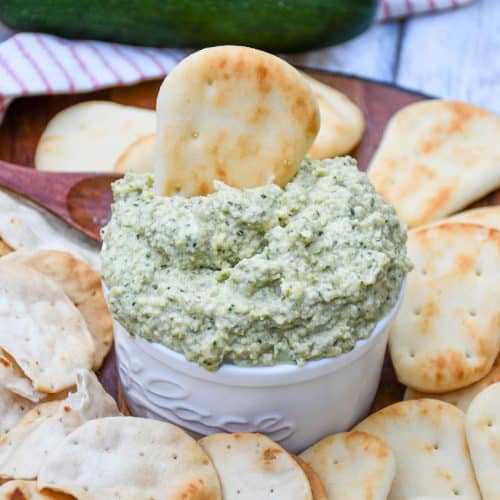 ZUCCHINI HUMMUS IN A SMALL WHITE BOWL SURROUNDED BY MINI NAAN BREAD AND PITA CRACKERS
