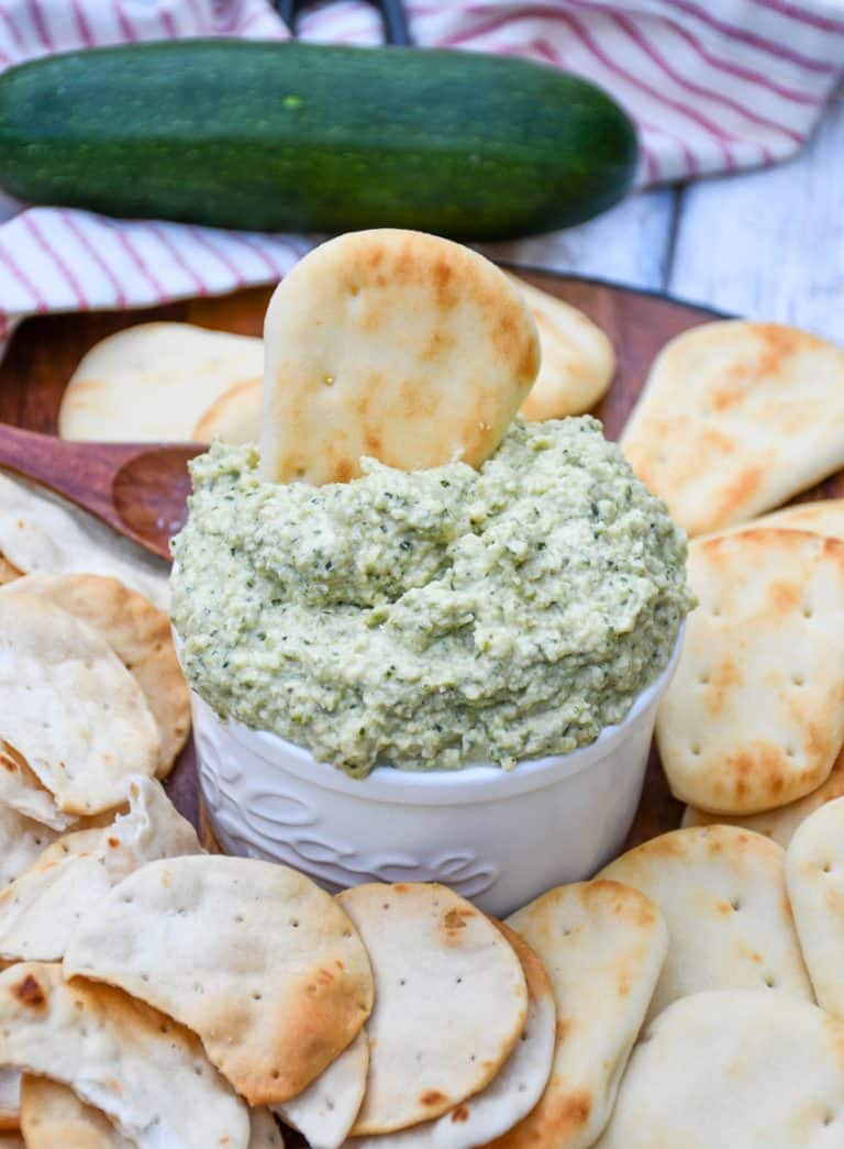 ZUCCHINI HUMMUS IN A SMALL WHITE BOWL SURROUNDED BY MINI NAAN BREAD AND PITA CRACKERS