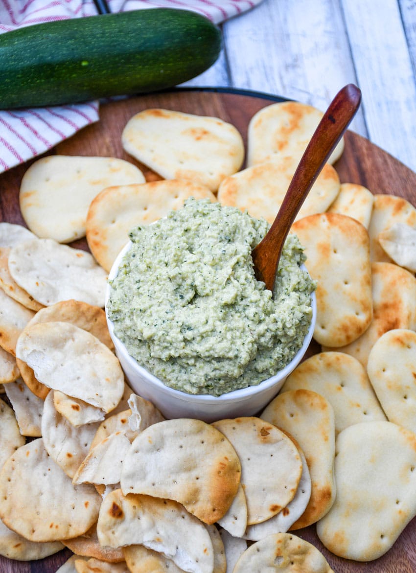 ZUCCHINI HUMMUS IN A SMALL WHITE BOWL SURROUNDED BY MINI NAAN BREAD AND PITA CRACKERS