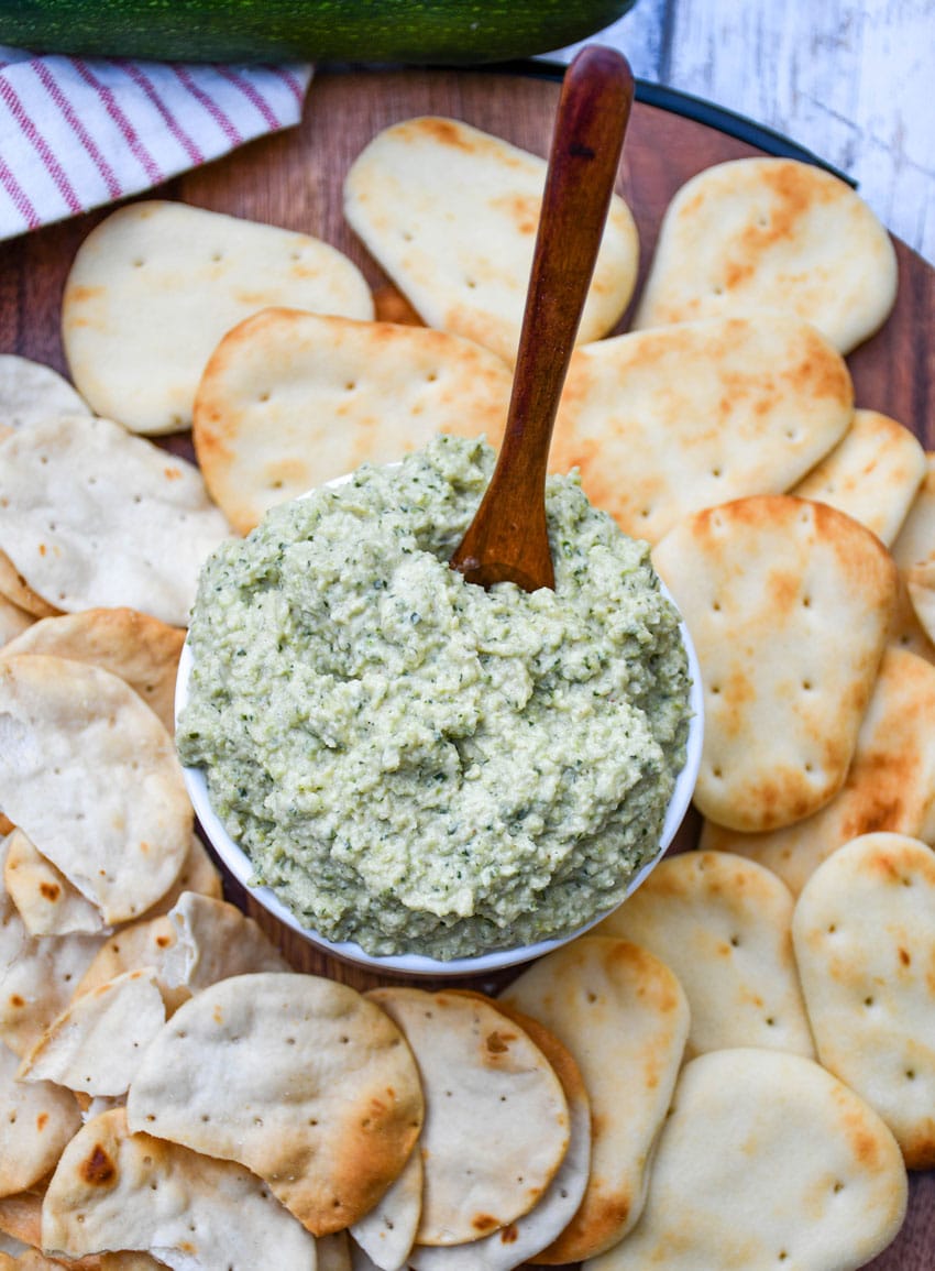 ZUCCHINI HUMMUS IN A SMALL WHITE BOWL SURROUNDED BY MINI NAAN BREAD AND PITA CRACKERS