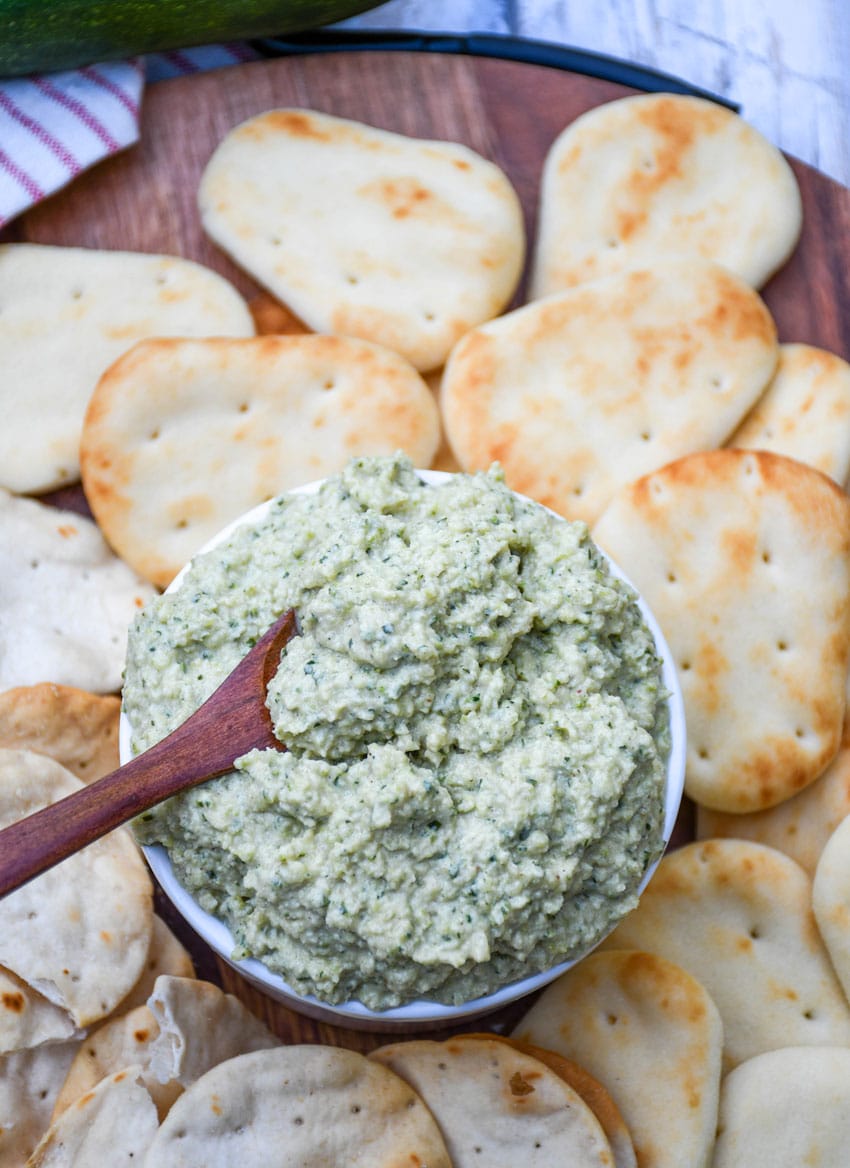 ZUCCHINI HUMMUS IN A SMALL WHITE BOWL SURROUNDED BY MINI NAAN BREAD AND PITA CRACKERS