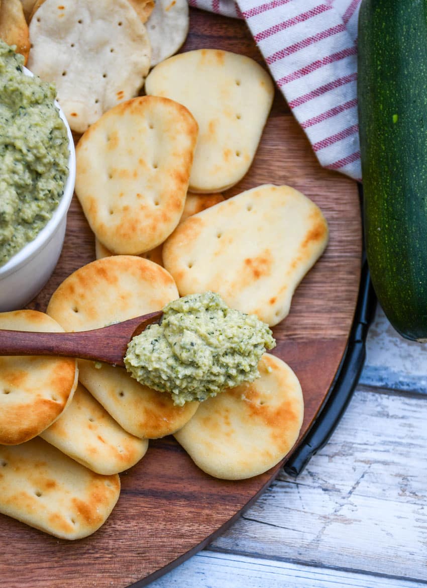a wooden spoon holding a scoop of homemade zucchini hummus resting on a wooden cutting board