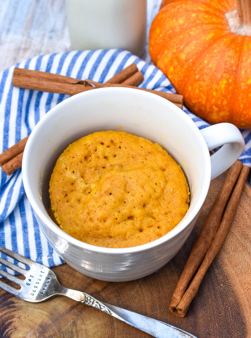 a microwave pumpkin mug cake on a wooden serving board with cinnamon sticks on the side