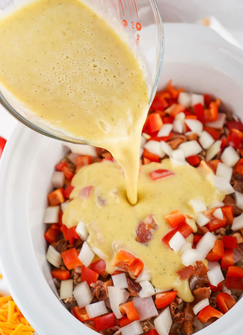 whisked eggs being poured over breakfast casserole ingredients in the white bowl of a slow cooker