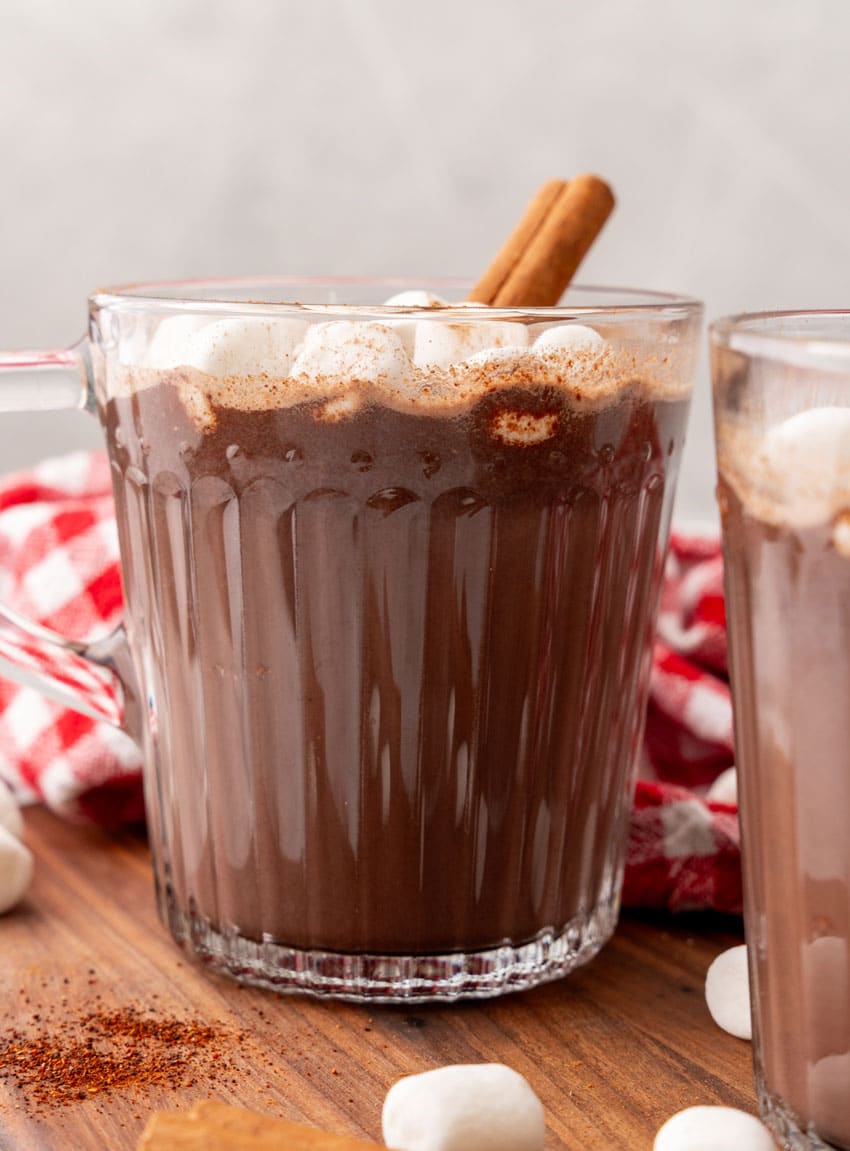 mexican hot chocolate studded with mini marshmallows and a cinnamon stick in two small glass jars on a wooden serving board
