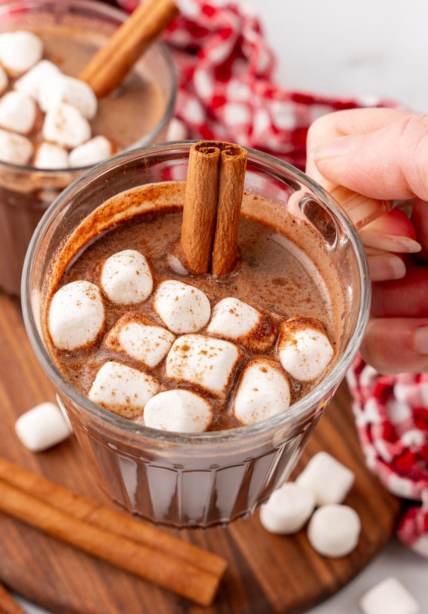 mexican hot chocolate studded with mini marshmallows and a cinnamon stick in two small glass jars on a wooden serving board