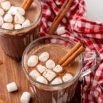 mexican hot chocolate studded with mini marshmallows and a cinnamon stick in two small glass jars on a wooden serving board