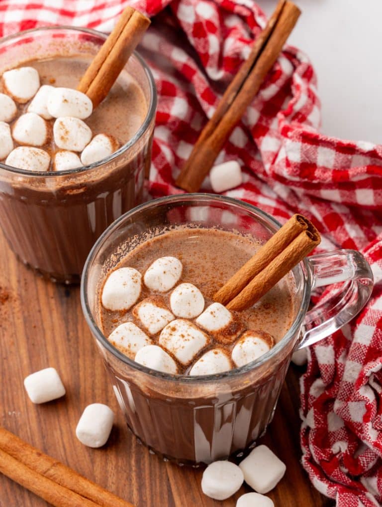 mexican hot chocolate studded with mini marshmallows and a cinnamon stick in two small glass jars on a wooden serving board