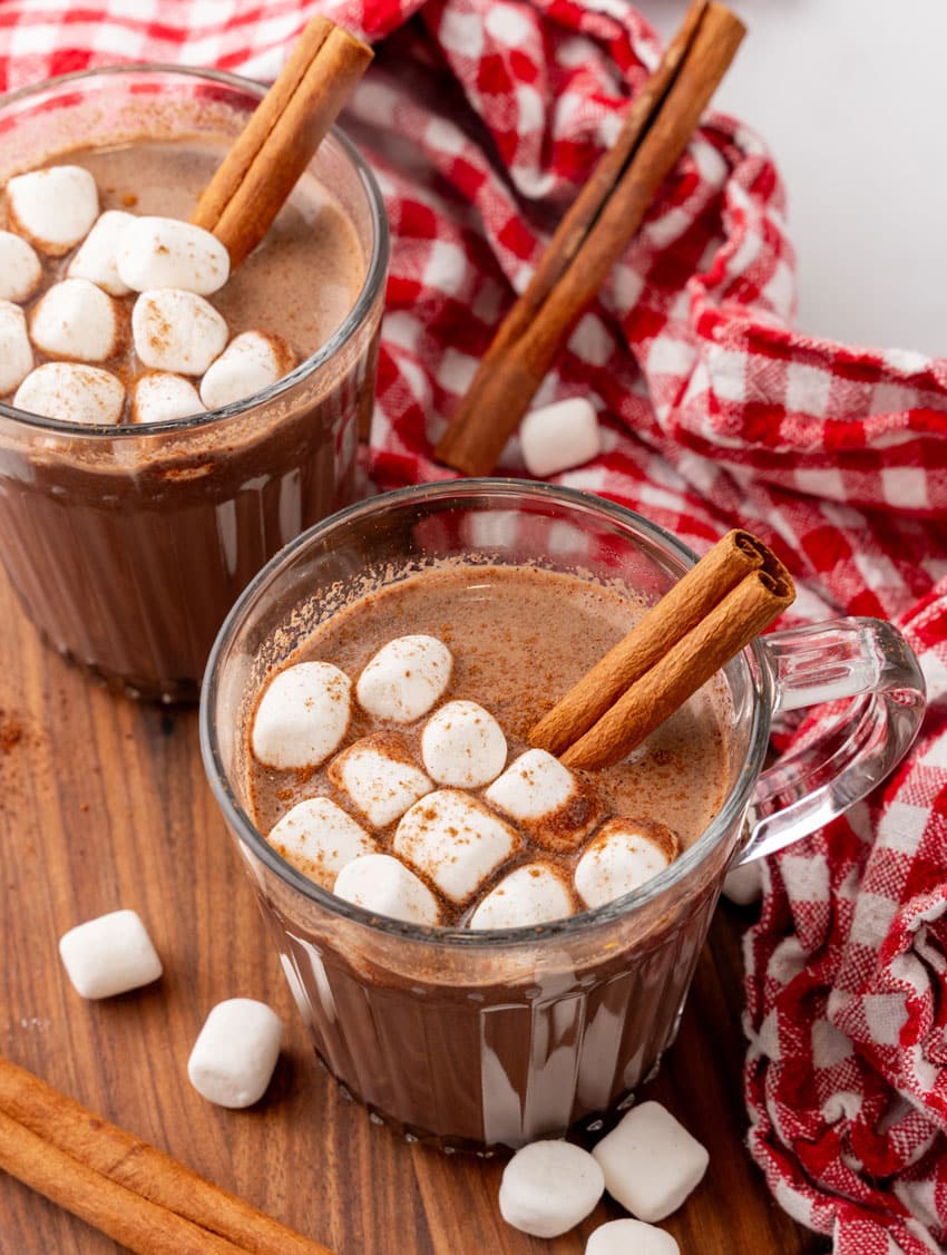 mexican hot chocolate studded with mini marshmallows and a cinnamon stick in two small glass jars on a wooden serving board
