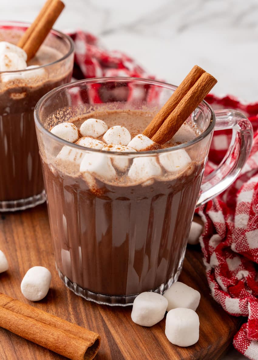 mexican hot chocolate studded with mini marshmallows and a cinnamon stick in two small glass jars on a wooden serving board