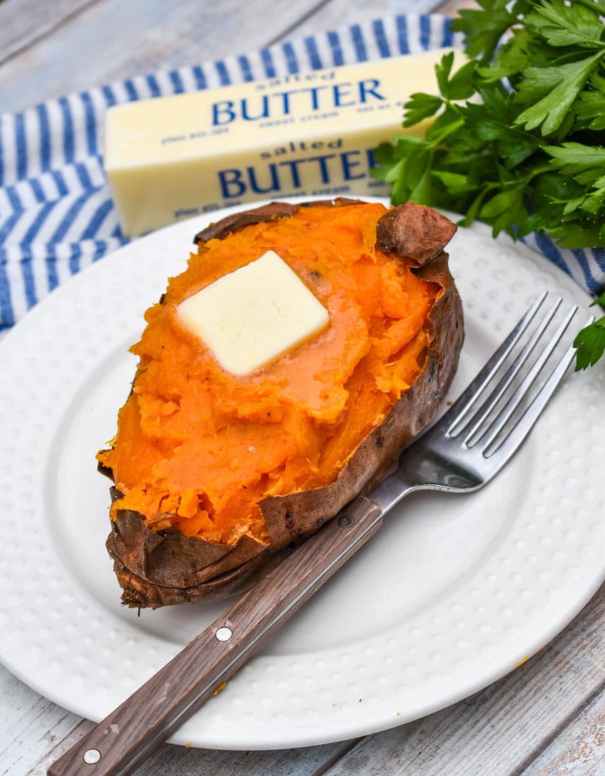 an air fryer baked sweet potato topped with a melting pat of butter on a small white plate with a fork resting on the side