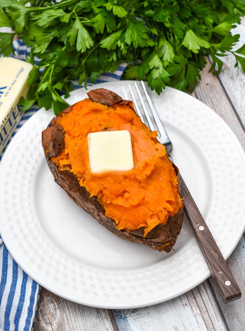 an air fryer baked sweet potato topped with a melting pat of butter on a small white plate with a fork resting on the side