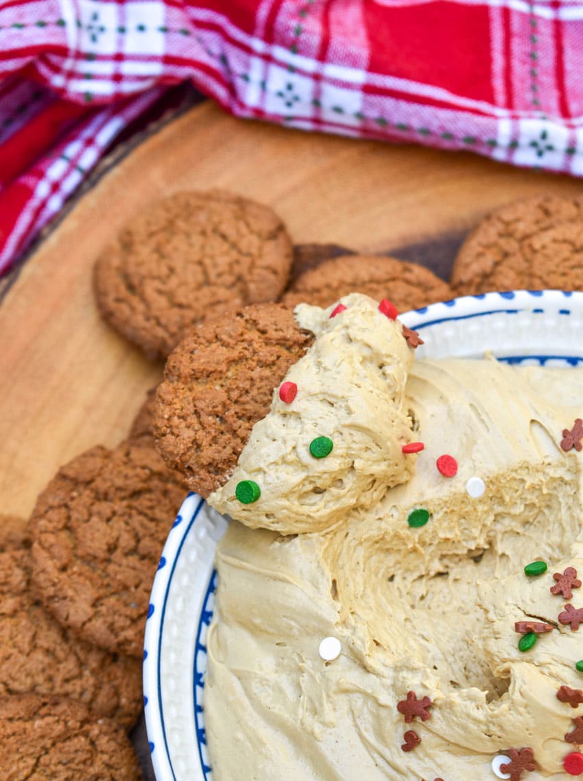 a gingersnap cookie topped with gingerbread cheesecake dip resting on the edge of a white serving bowl