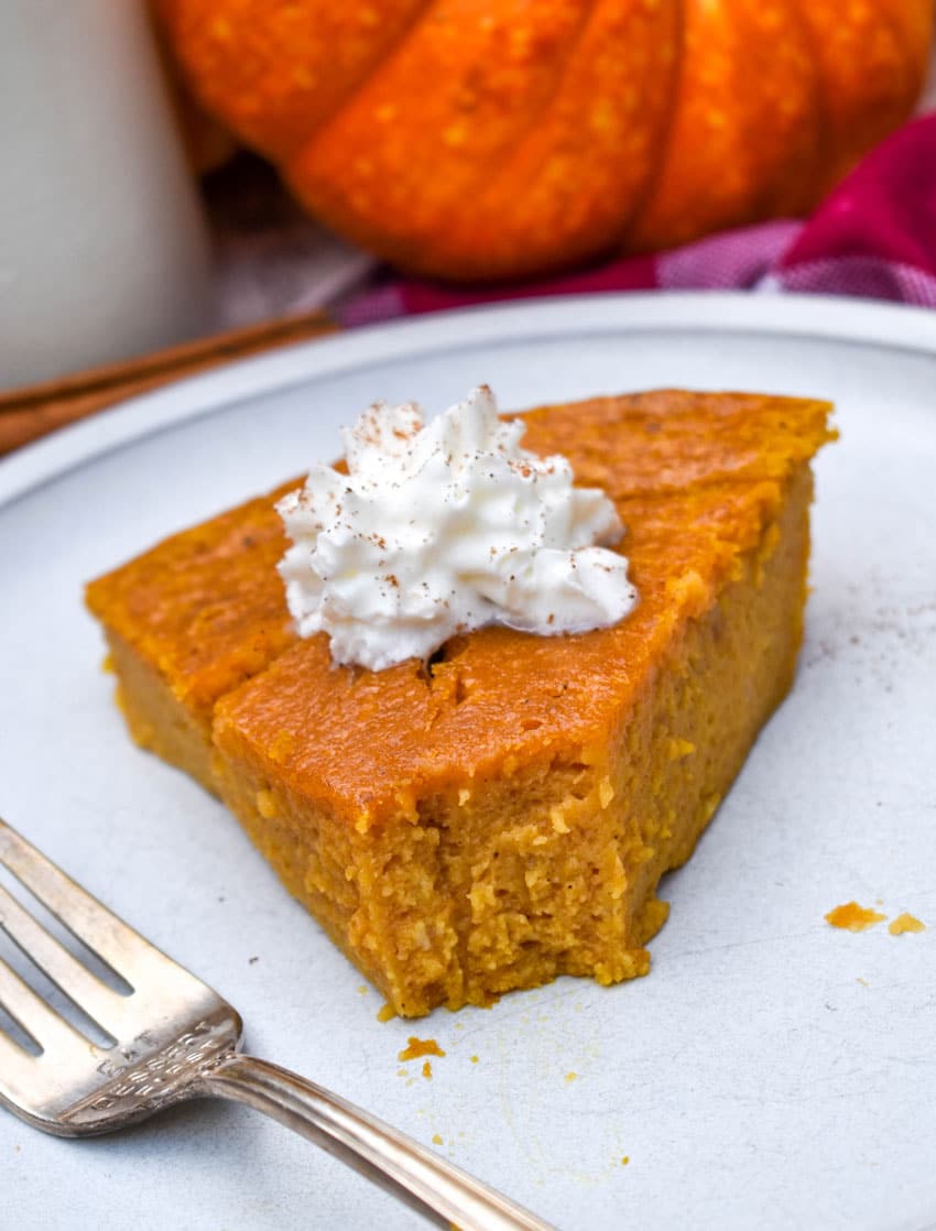 a sliced of impossible pumpkin pie topped with a dollop of whipped cream on a white plate with a silver fork resting on the side