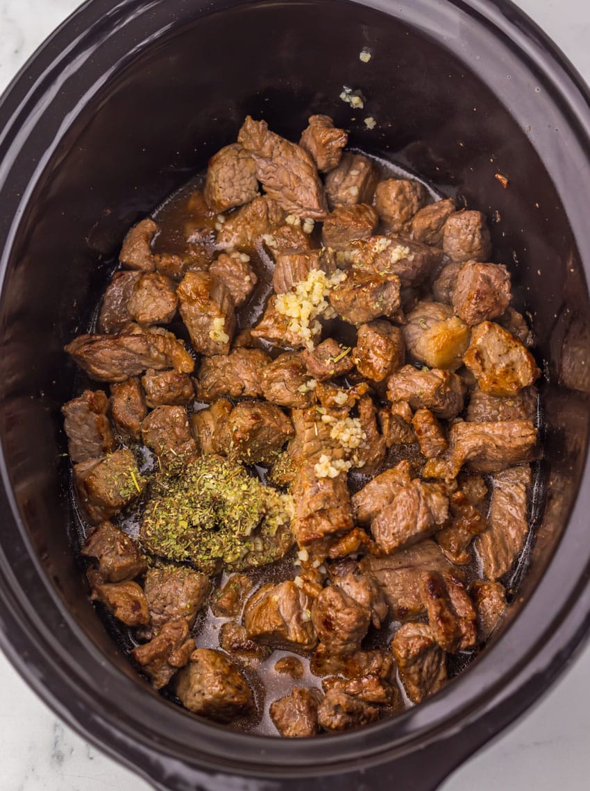 seared steak, minced garlic, and seasonings in the black bowl of a slow cooker