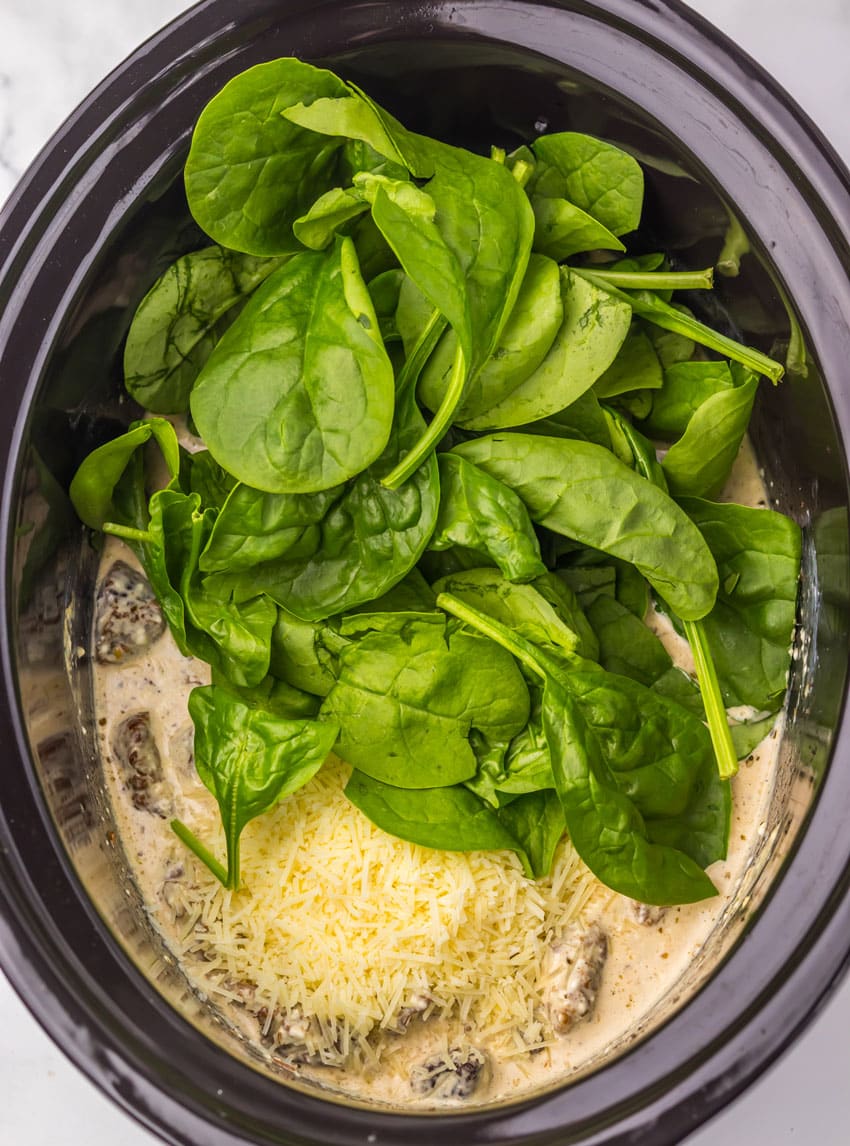 fresh spinach leaves, shredded Parmesan cheese, and cream over steak in the black bowl of a slow cooker